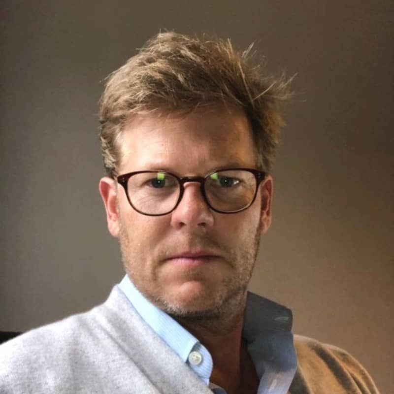 A man with light brown hair and glasses is looking directly at the camera. He is wearing a light blue shirt under a gray sweater. The background is softly lit, creating a neutral tone that complements his serious expression.