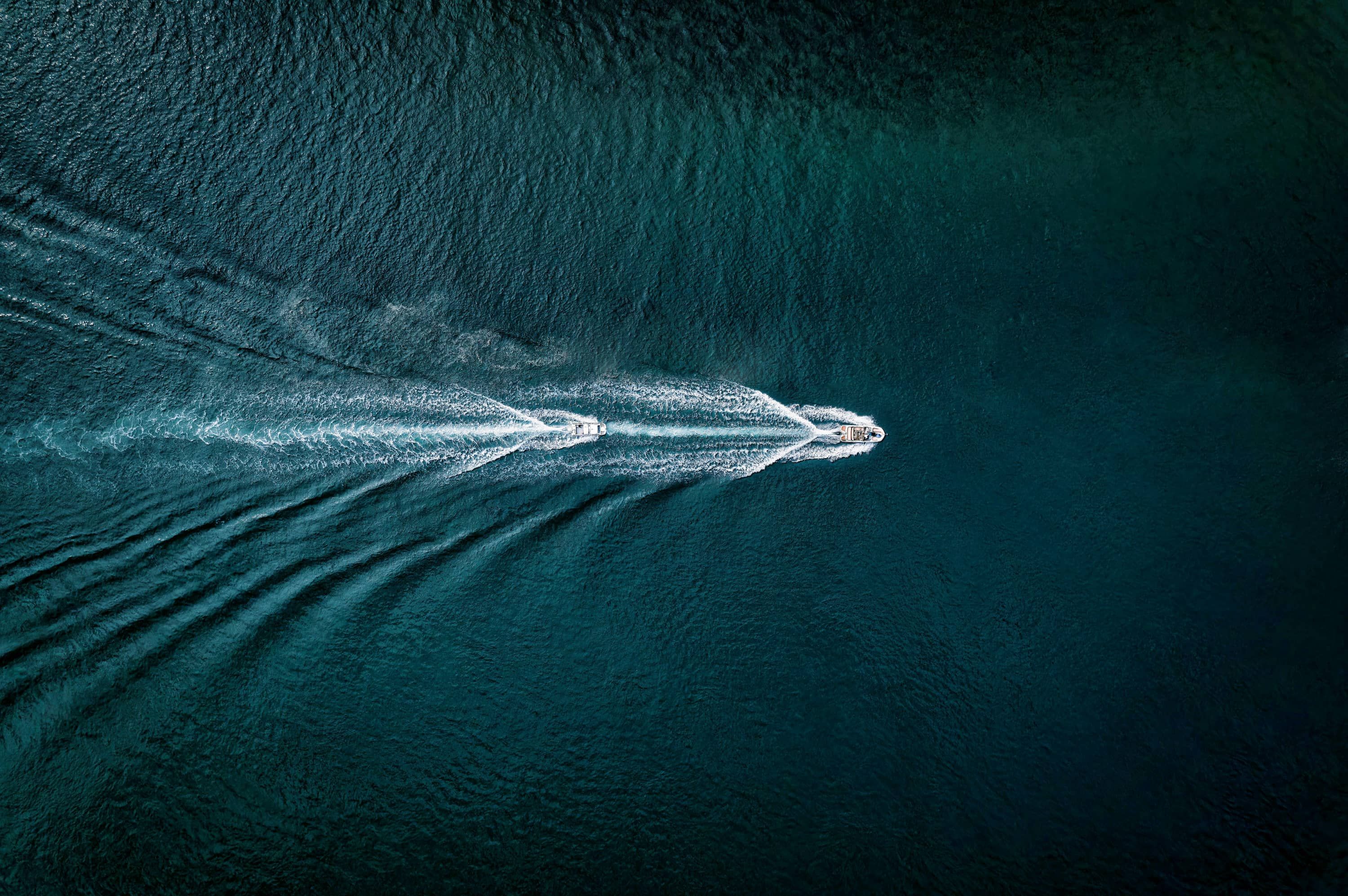 Aerial view of two boats traveling on dark blue water, leaving white wake trails behind them. The scene captures the patterns created on the water's surface as the boats move in different directions.